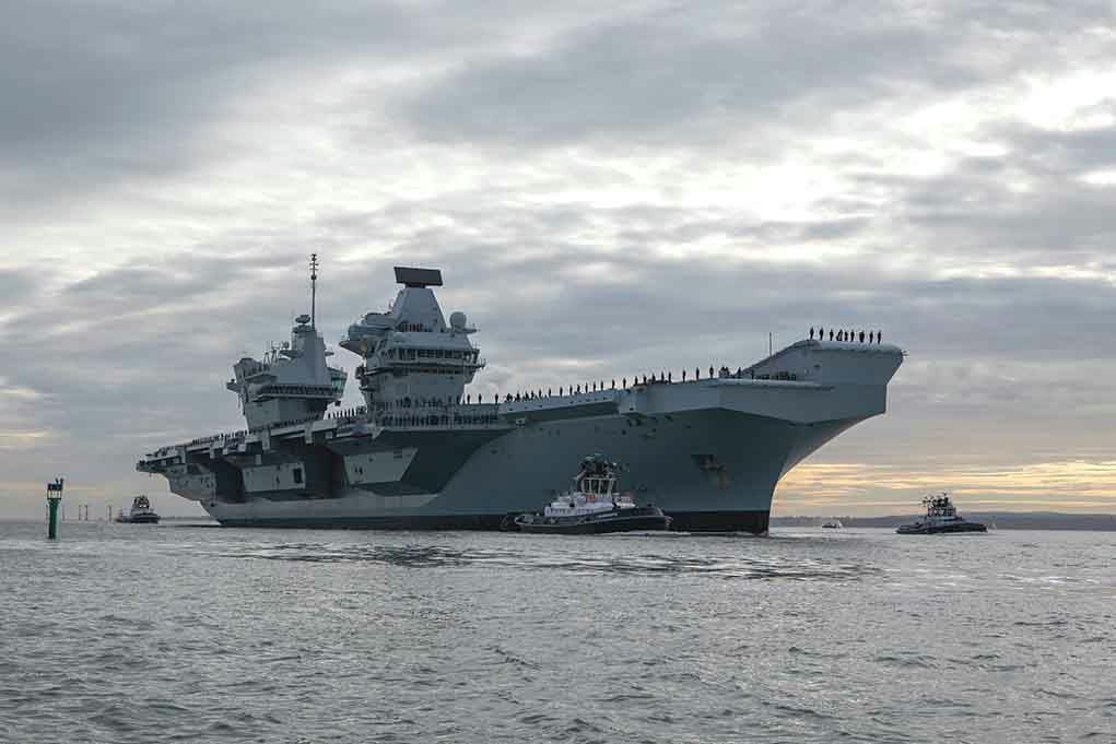 Aircraft carrier with tugboats on a cloudy sea.