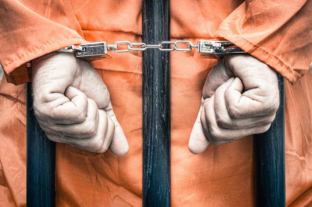 Handcuffed person in orange jumpsuit behind bars.