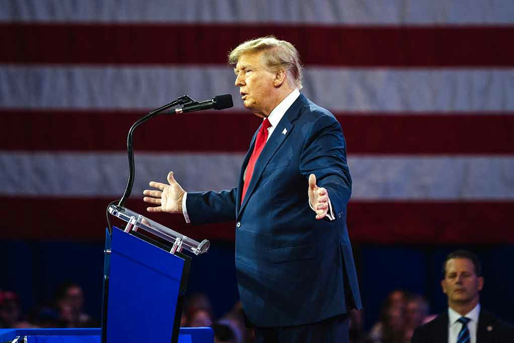 Man speaking at podium with American flag backdrop.