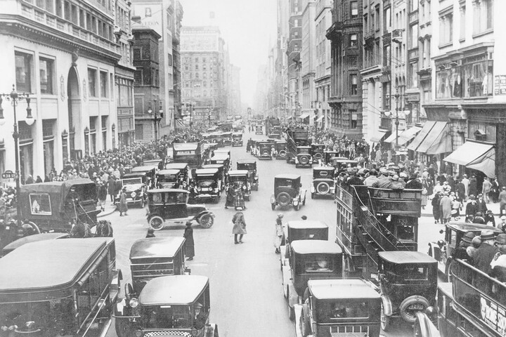 A busy city street from 100 years ago filled with vintage cars and crowds of people