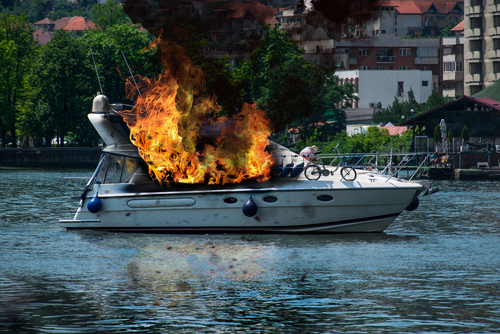 Boat on fire on a river; buildings in background.