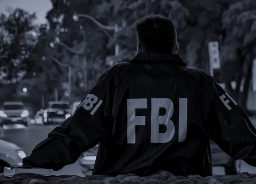 Person in FBI jacket stands, backs to camera, outdoors.
