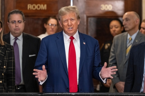 A public figure speaking at a press conference with a group of people in the background